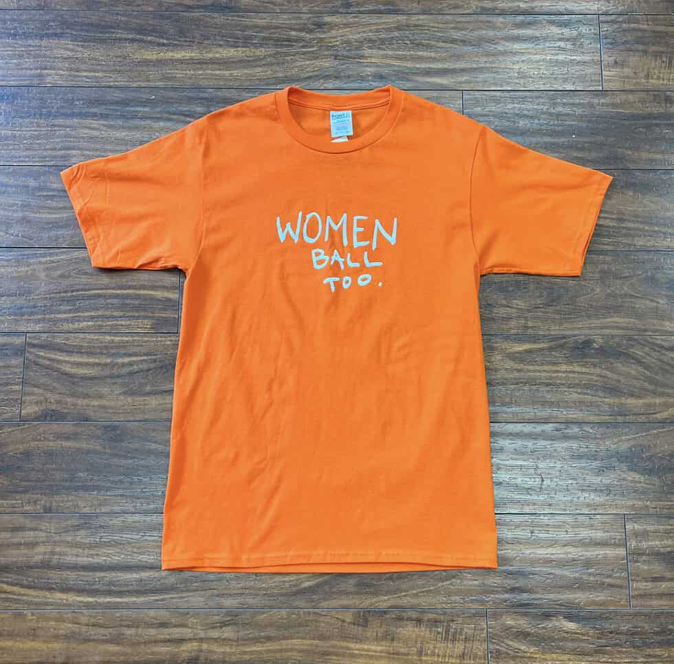 Orange "Women Ball Too" t-shirt laid flat on a wooden floor, size small.